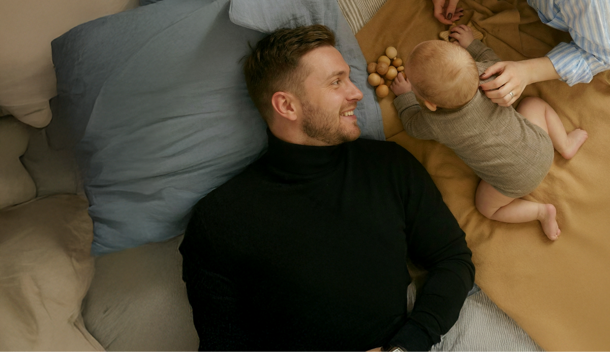 Man lying on a bed with a baby and another person in a home setting